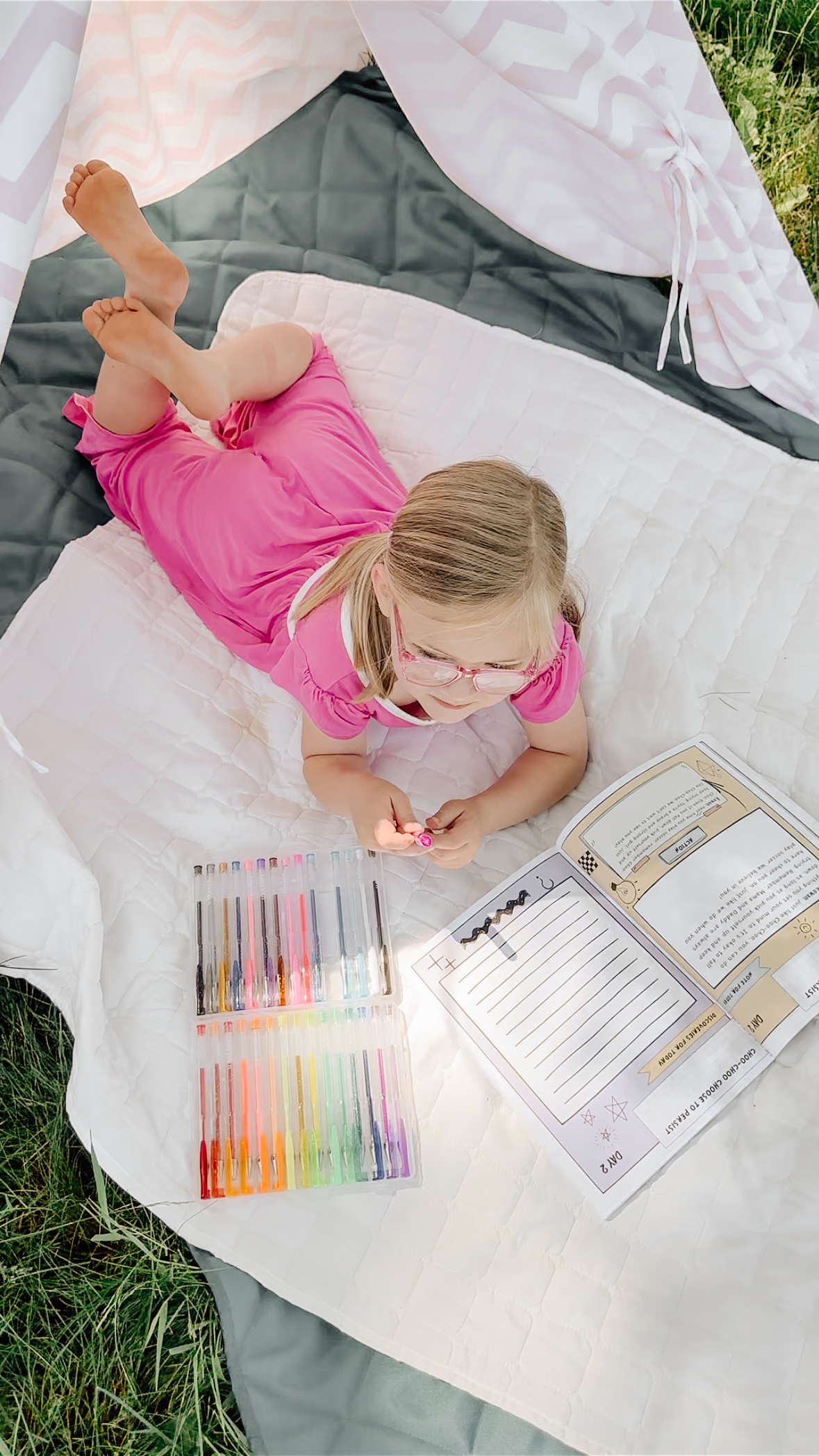 Girl on blanket under teepee with colorful pens and open book